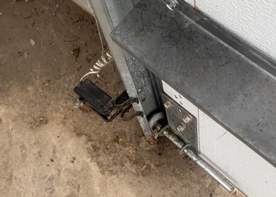 Home garage door repair in Los Angeles, CA, showing lower track hardware, bottom bracket, roller, and loose safety sensor wiring.